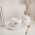 Biały zestaw porcelanowy z personalizacją - imbryk i filiżanka ze spodkiem z imionami dzieci dla babci na każdą okazję Zbliżenie na złote zdobienie na białym filiżance porcelanowej imbryk i filiżanka z imionami dzieci na spodku Nadzwyczajnie.pl